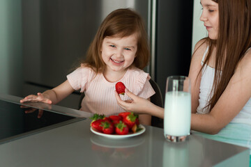 Two girls sisters sitting at kitchen, eating strawberry, drinking milk