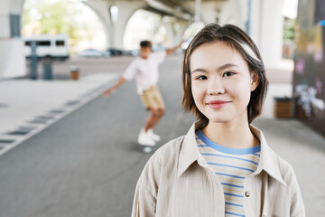 Portrait of young teenage girl in skatepark looking at camera and smiling with boy riding skateboard in background, copy space