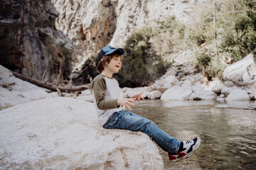 Caucasian school boy sits on a riverside rock in the canyon with mountain cliffs in the background. Kid child taking a rest on a boulder near mountain river bank. © Anna