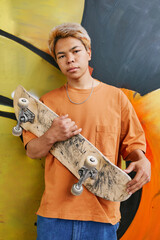 Colorful portrait of young teenage boy with skateboard standing against graffiti wall in orange and looking at camera - obrazy, fototapety, plakaty