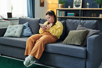 Obraz premium Portrait of modern girl with Down syndrome sitting on sofa in living room at home using digital tablet