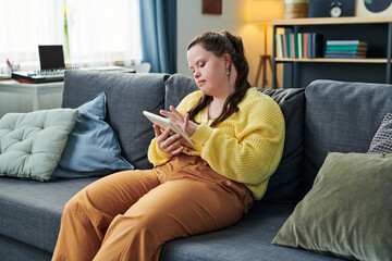 Young woman with disability spending time at home sitting on couch in living room scrolling news...
