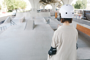 Back view of young teenage girl wearing protective guards and helmet in skating park outdoors, copy space