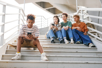 Fototapeta premium Full length portrait of teenage boy using smartphone outdoors while sitting on metal stairs with group of friends in background, copy space