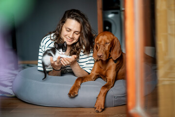 Portrait of a young woman with a Hungarian Pointer dog and a small kitten in her arms lying at home in a room on a bag chair