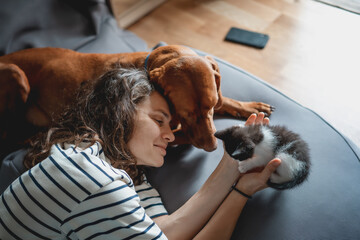 Portrait of a young woman with a Hungarian Pointer dog and a small kitten in her arms lying at home in a room on a bag chair - 506482610