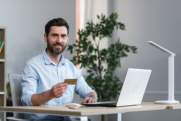 Happy businessman makes online purchases working in the office, husband holds credit bank card and uses laptop