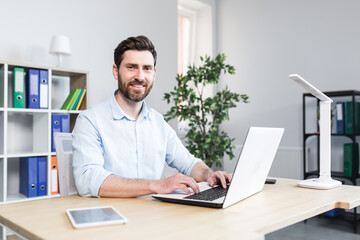 Happy businessman working in bright modern office behind laptop man with beard smiling and looking...
