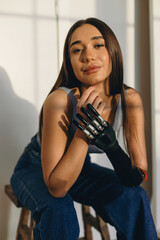 Gorgeous self-confident youthful woman with prosthetic hand instead of amputated arm after injury sitting on stool, bending to camera, putting head high, keeping elbows on knees, wearing jeans and top