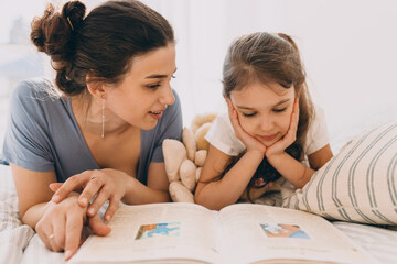 Loving and caring young Caucasian mother reading fairy tale to her preschool daughter, lying in bed in front of opened book, girl listening attentively, looking at pictures, holding her head in hands