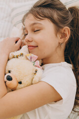 Vertical picture of adorable little Caucasian girl of 8-years-old in white t-shirt or pajama sleeping with her teddy bear toy in bed, smiling having sweet dreams. Happy carefree childhood