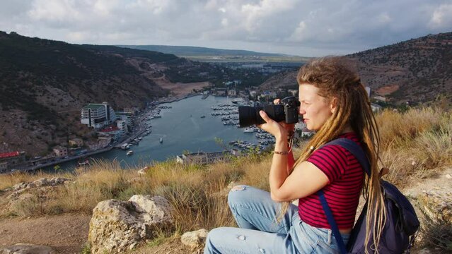 Attractive girl with dreadlocks takes pictures of river in gorge with ships and buildings on shore. Profile side view. Young woman photographer shoot photos of water landscape with mountains