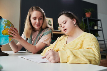 Young female teacher sitting at table next to her student with Down syndrome helping with task...