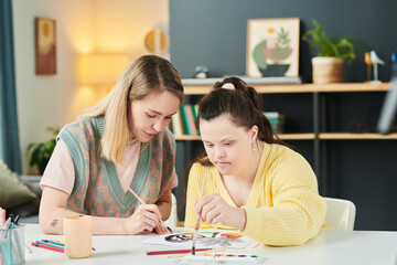 Modern girl with Down syndrome painting picture with watercolors during individual art class with young female teacher