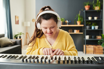 Portrait of joyful young Caucasian girl with Down syndrome wearing headphones spending time at home learning to play electronic keyboard