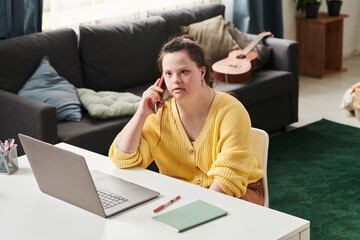 Modern young woman with Down syndrome sitting at table in front of laptop speaking on phone while...