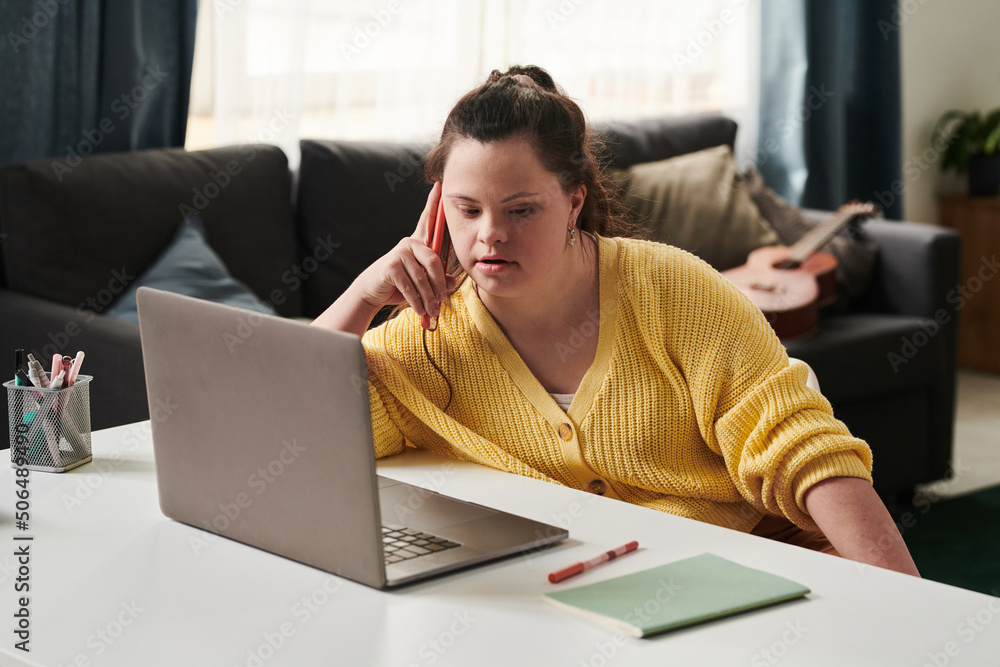 Wall mural young woman with down syndrome sitting relaxed at table at home with laptop on it talking on phone w