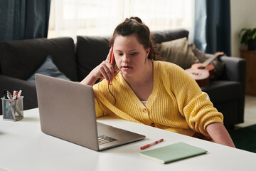 Young Woman with Down syndrome sitting relaxed at table at home with laptop on it talking on phone...
