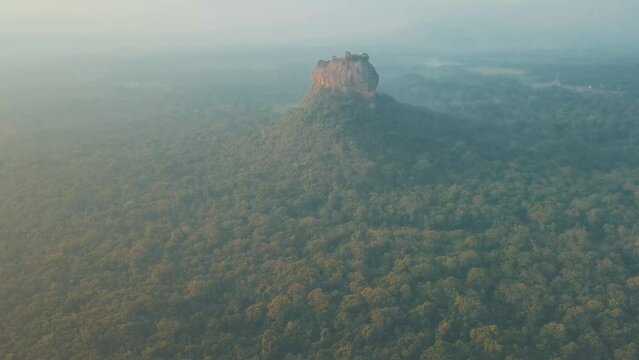 Aerial drone shot of Sigiriya Lion Rock in Sri Lanka at sunrise with light mist and haze filled skies overlooking natural tropical forest and lush mountain ranges with orange blue tones