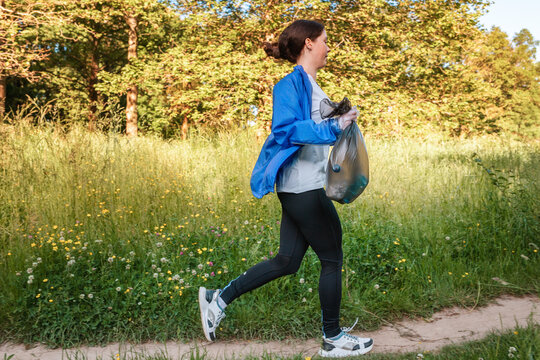 Young woman in sportswear jogging at the park and holding a bag of garbage. Side view. The concept of plogging, environment protection and volunteering