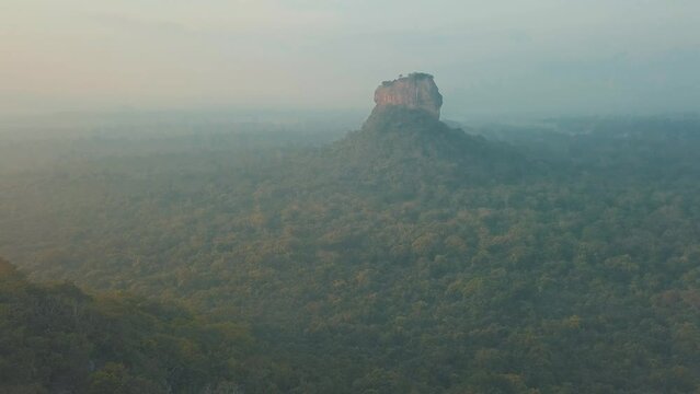 Aerial drone shot of Sigiriya Lion Rock in Sri Lanka at sunrise with light mist and haze filled skies overlooking natural tropical forest and lush mountain ranges with orange blue tones