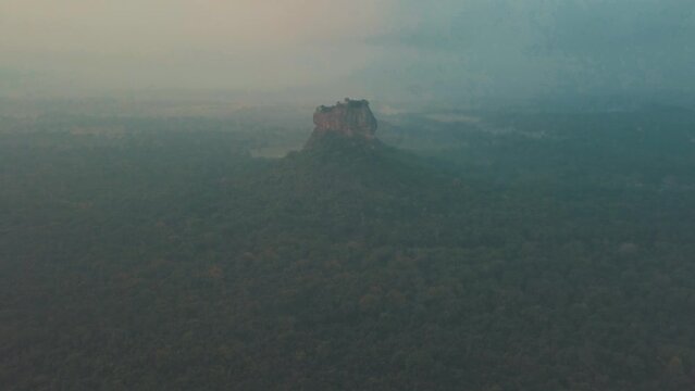 Drone panning point of interest Sigiriya Lion Rock in Sri Lanka at sunrise with light mist and haze filled skies overlooking natural tropical forest and lush mountain ranges with gray and blue tones