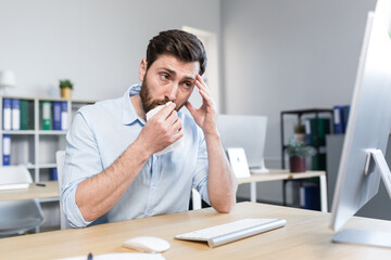 Sick and tired young man at work. Office worker, manager, freelancer sitting at a desk in the office does not feel well, wipes his nose, has a runny nose, coughs, headaches, holds his head.