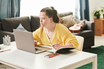 Portrait of young female university student with Down syndrome sitting at table at home using...