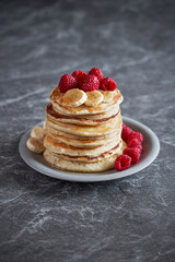 American pancakes with raspberries and bananas over dark minimal background. Delicious summer breakfast on black stone table. Minimal design.