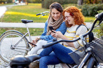Two friends woman sitting on the bench and looking at mobile phone and smile
