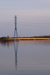 Tall electricity pylon casting reflection on the calm water during the sunset.