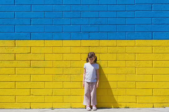 little toddler girl sad on the background of a brick wall painted in the colors of the Ukrainian flag, the concept of supporting the children of Ukraine during the war
