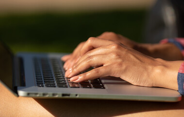Close-up shot of a young woman typing on a laptop keyboard while sitting on grass. 