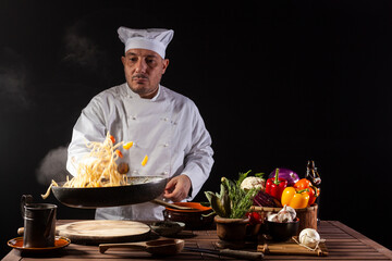 Male chef in white uniform holding a frying pan, sautéing spaghetti with fresh vegetables flying in the air