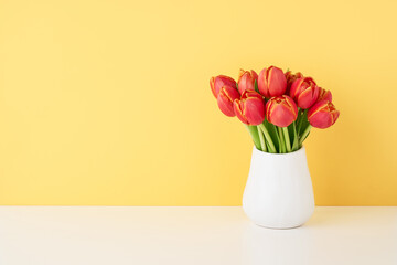 Bunch of red tulips in a white ceramic vase on the white table by the wall. Copy space for text