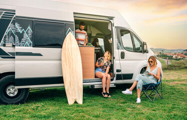 Group of friends traveling in a camper van. Two women sitting in front of camper van while young...