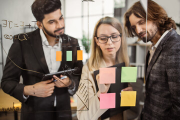 Caucasian female manager making sticky notes on glass board and sharing ideas with multiethnic male team during presentation. Creative colleagues using tablet and clipboard at modern coworking space.