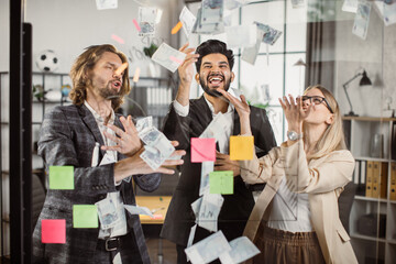 Happy multicultural business people in formal wear laughing and throwing out banknotes during meeting in modern office. Financial success and cooperation concept.