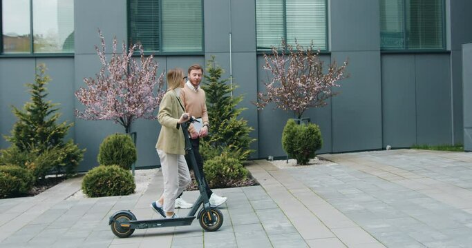 Caucasian young woman and man walking on street with kick e-scooter and laptop talking and laughing by modern office buildings. Technology, partnership, outside