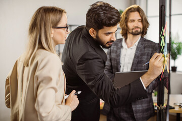 Handsome indian man adding data with marker on glass wall during meeting with colleagues at office. Multicultural project group preparing new ideas for developing business.