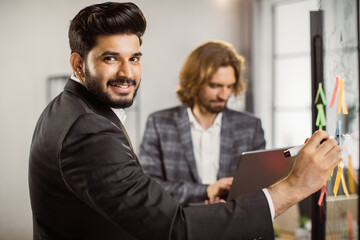 Successful indian businessman smiling and looking at camera while drawing business plan on glass office board. Caucasian male colleague standing near with portable laptop in hands.