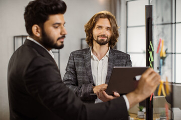 Positive caucasian man in suit holding laptop and looking at camera while his indian business partner writing on glass board. Multi ethnic workmates planning on new strategies in office.