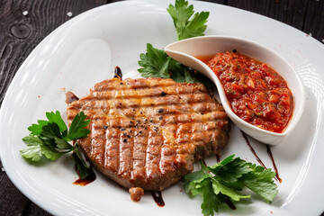 Pork meat steak under spicy salsa sauce with greens on a white plate. 