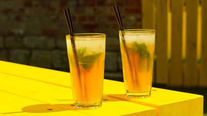 Two tall glasses of cold mint lemonade with black plastic straws stand on a yellow wooden table. Summer drinks theme on a hot day