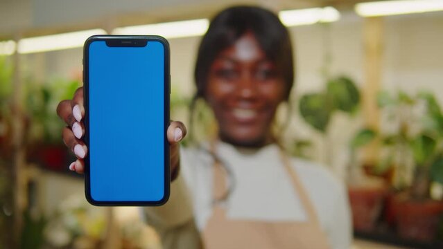 Forist African American woman holding phone with chroma key. Greenhouse worker female botanist smiling, small business owner. Gardener working in flower shop, plant store. 
