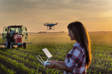 Farmer woman driving drone in field with tractor in background - obrazy, fototapety, plakaty