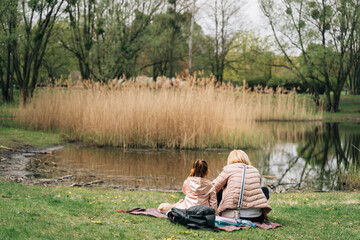 Anonymous mother with little daughter sitting at lakeside during picnic in park