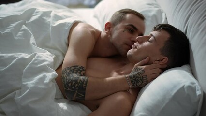 Side view young men sleeping in bed together waking up in the morning kissing cheek. Happy loving Caucasian gay couple in bedroom at home indoors. Leisure and love concept