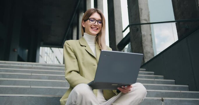 Good -looking young caucasian woman in glasses wearing a green jacket sitting with crossing legs on the stairs outside, typing on her laptop with satisfied face