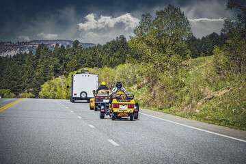 Two three wheel motorcycles with luggage follow camper van on four lane highway toward stormy...